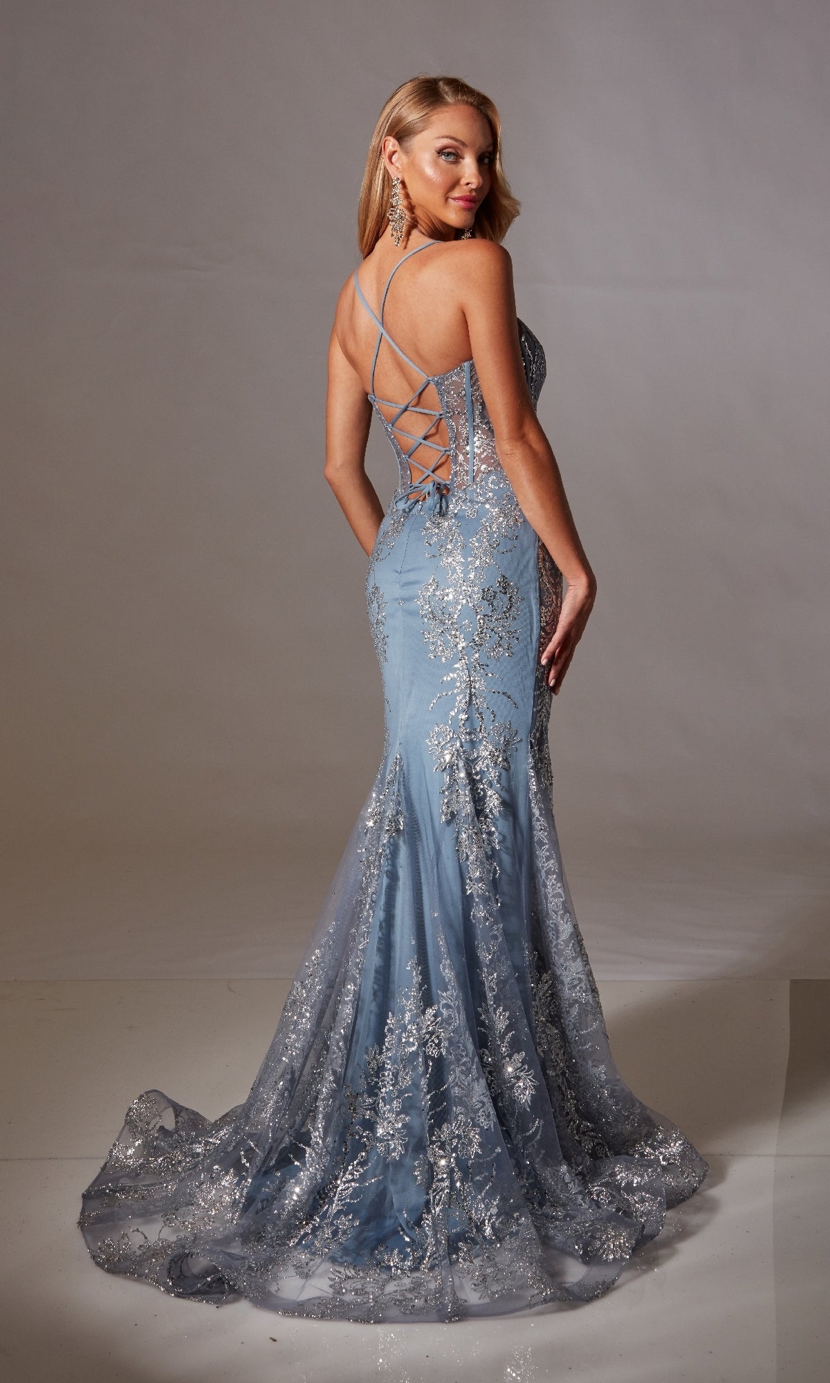A woman poses in the Aerie Couture 251, a long glitter-print prom dress featuring a light blue mermaid silhouette, silver embellishments, and a lace-up corset bodice with an open back design.